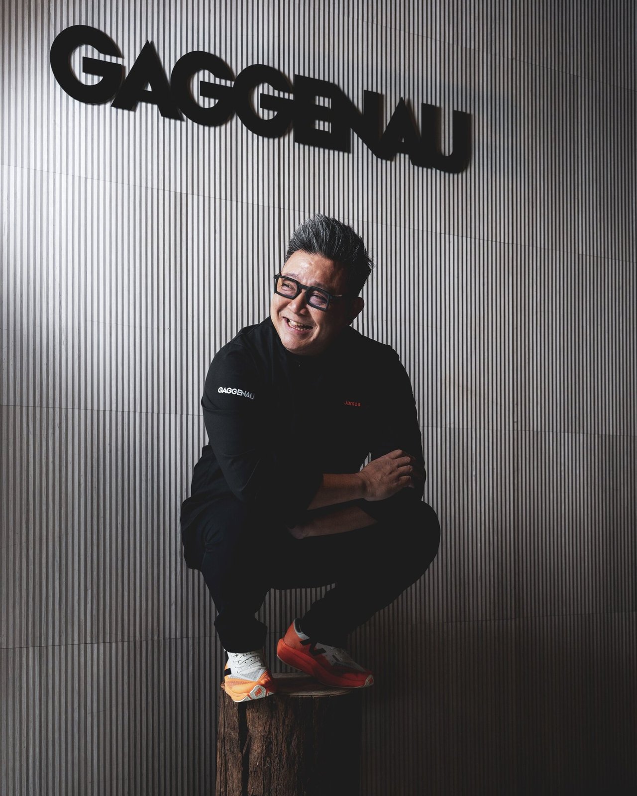 James Won seated on the wooden stump in front of the GAGGENAU sign &mdash; tighter frame focusing on the figure and brand mark.