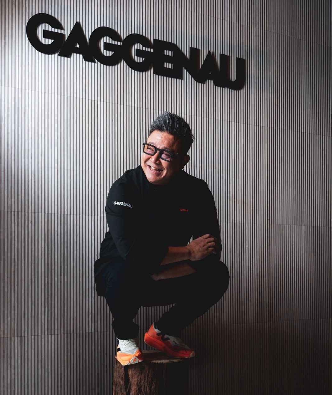 James Won on a wooden stump in front of the GAGGENAU sign &mdash; wider frame showing the full Inspiration Haus wall, orange Asics sneakers visible.