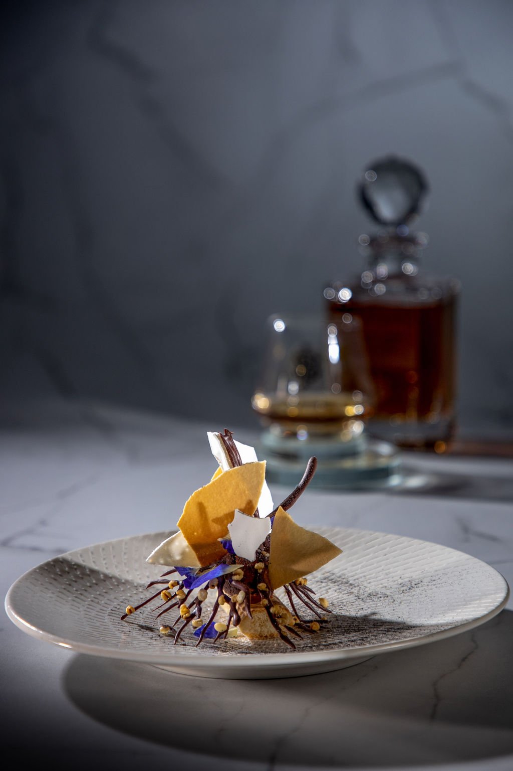 Discover Rarity dessert — Semai single-origin cocoa, Job's Tears, coconut crème tuile, pea-flower dust, on a textured plate; Johnnie Walker decanter behind.