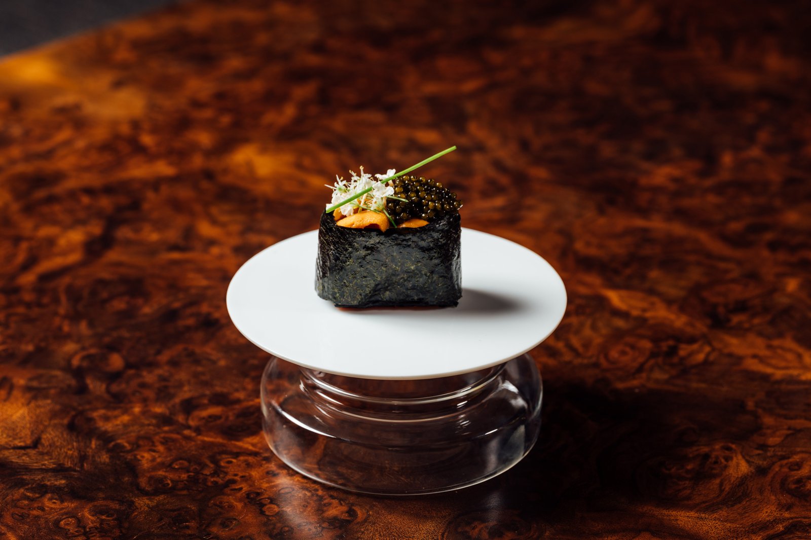 The James Won Custom Caviar served as a single nori-wrapped bite on a white pedestal plate against a dark burlwood table at Shin'Labo.
