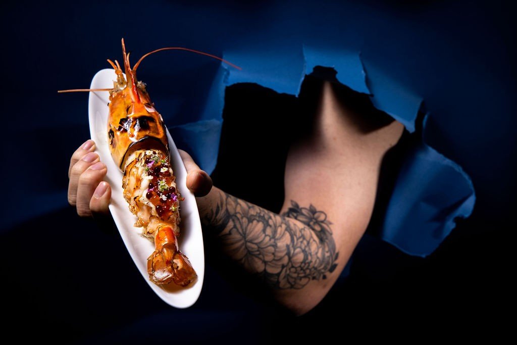 A whole glazed langoustine on a white oval plate, a second frame, held through torn dark-blue paper.