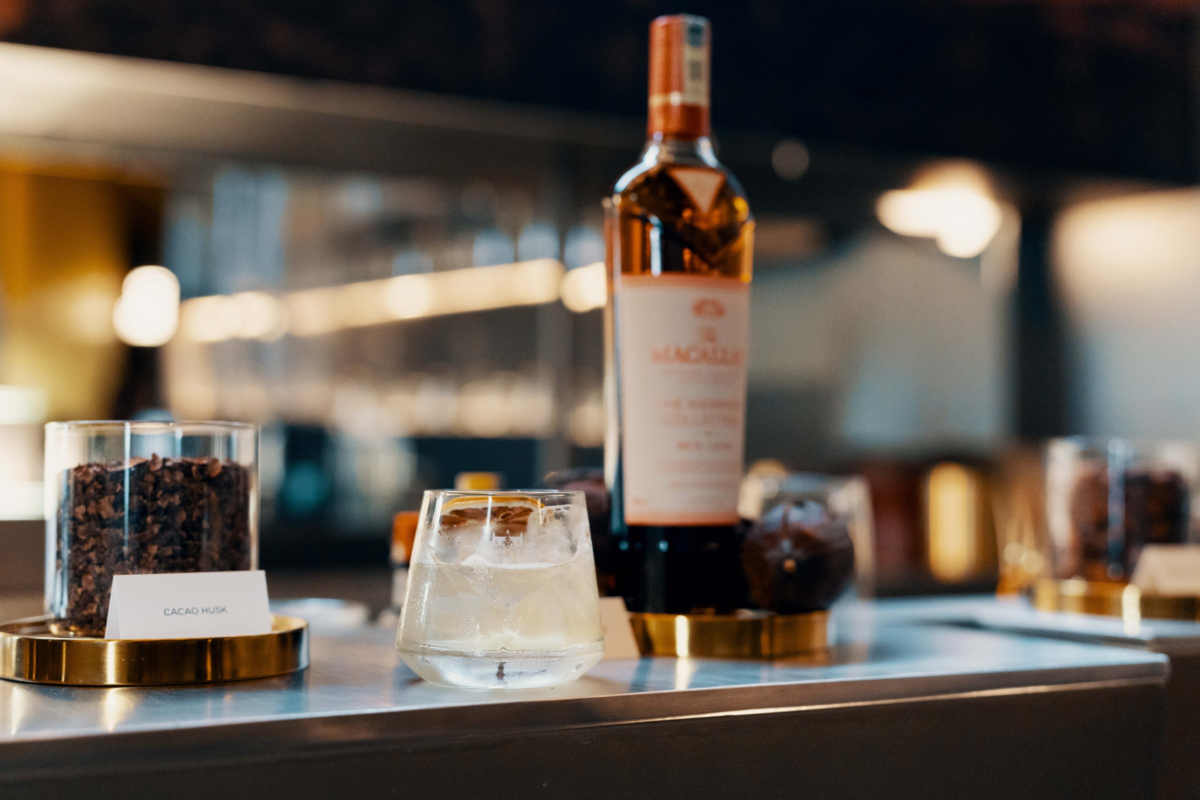 A bottle of The Macallan Rich Cacao on the bar beside jars of cacao and a glass, the room soft behind.