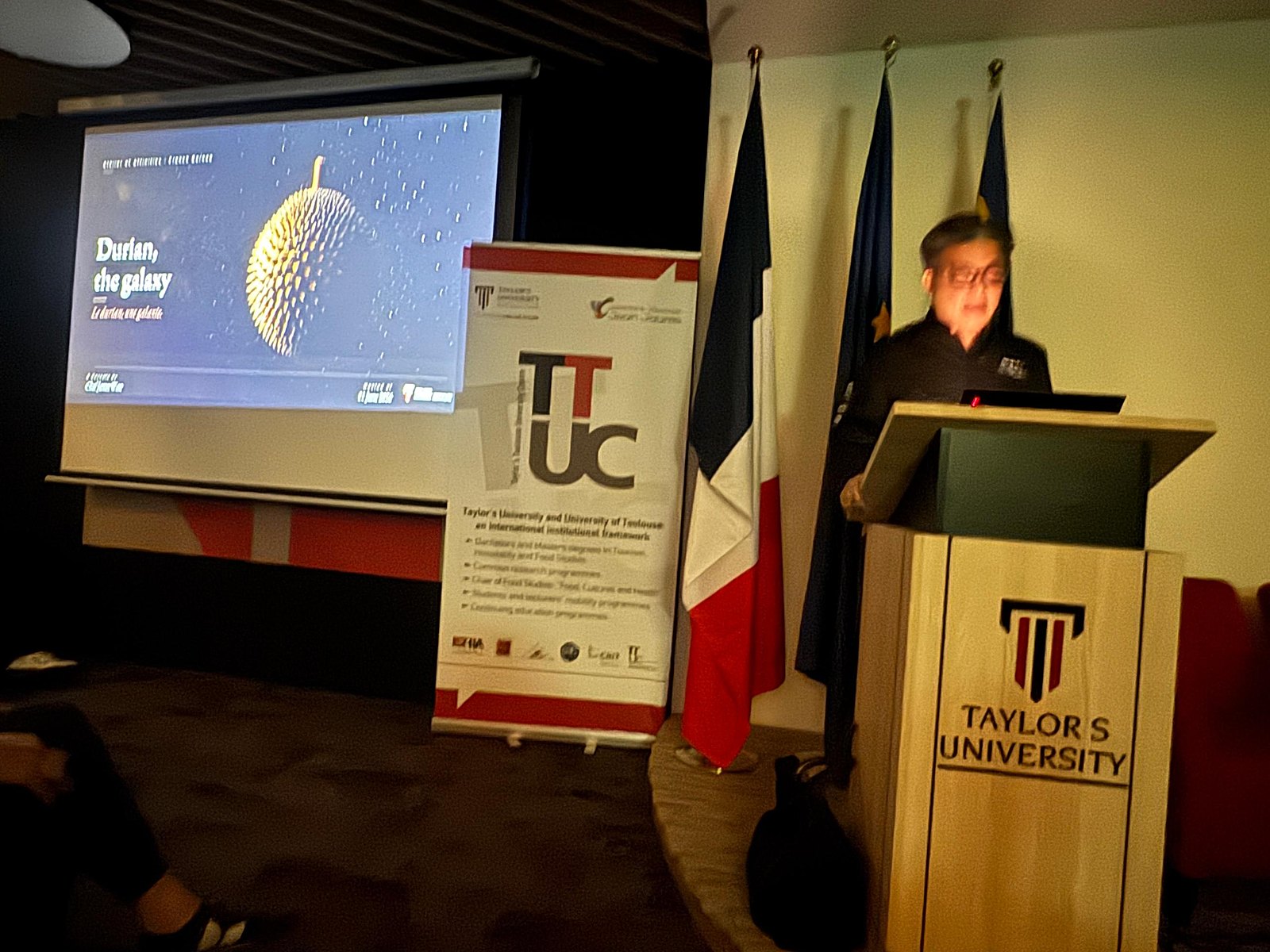 Chef James Won presenting Durian, the galaxy at the keynote at Taylor's University, French and Malaysian flags beside the lectern