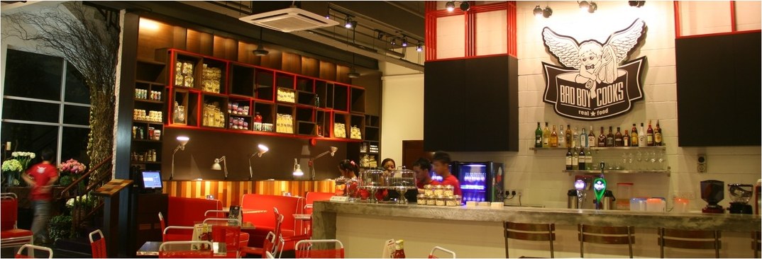 The BadBoyCooks Diner bar: a concrete counter with stools, the winged cherub logo on the wall above, bottles on a shelf, red pantry shelving to the left.
