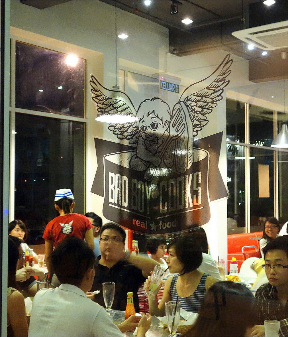 Diners at a full table in the evening, seen through the BadBoyCooks cherub logo on the window glass, with a server in a red T-shirt and paper cap.