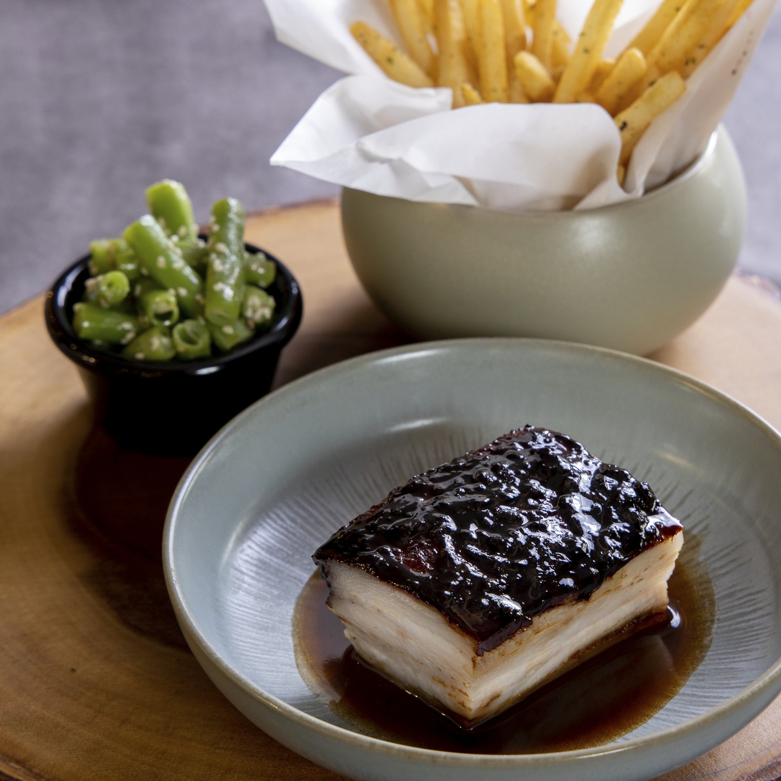 Bouchon Enfin glazed pork belly — square cut with sticky dark glaze, sesame green beans, and shoestring fries in a paper-lined cup.
