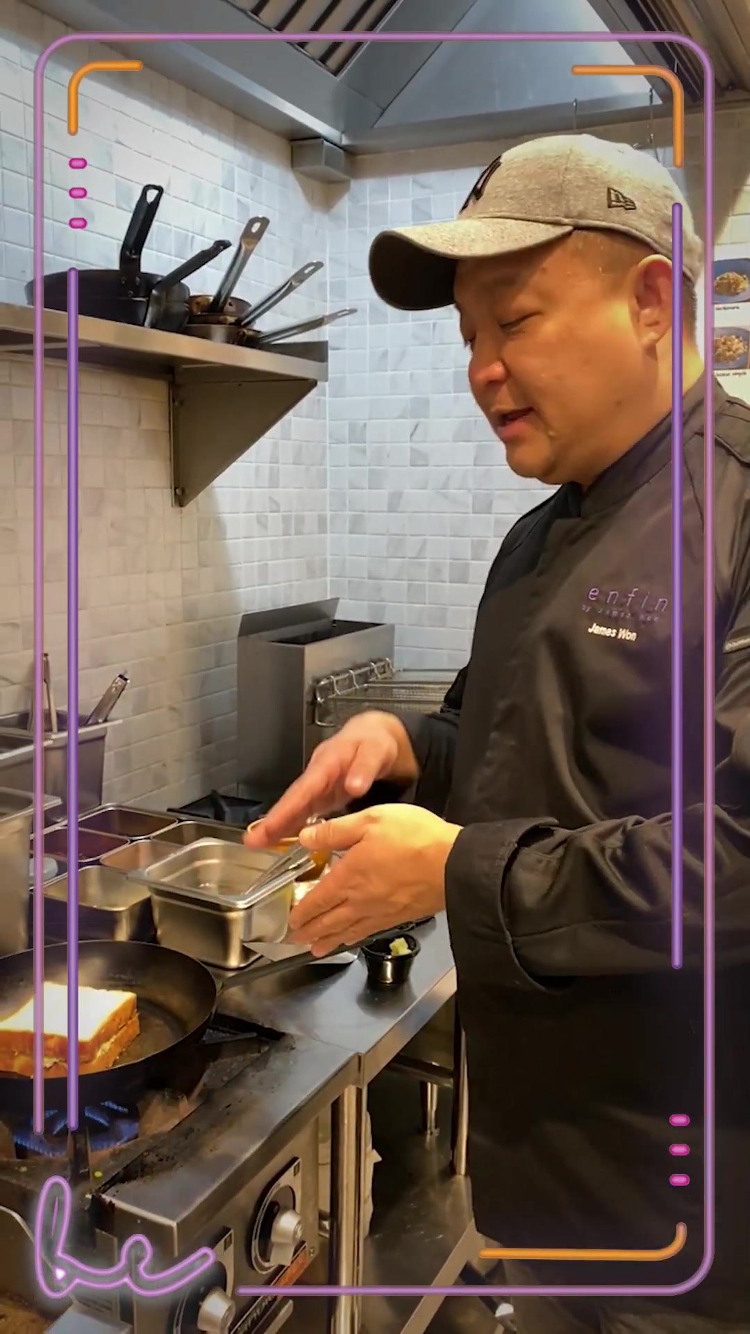 James Won at the stove in the Bouchon Enfin kitchen, a grilled cheese sandwich toasting in a black pan, pans racked on the tiled wall behind.