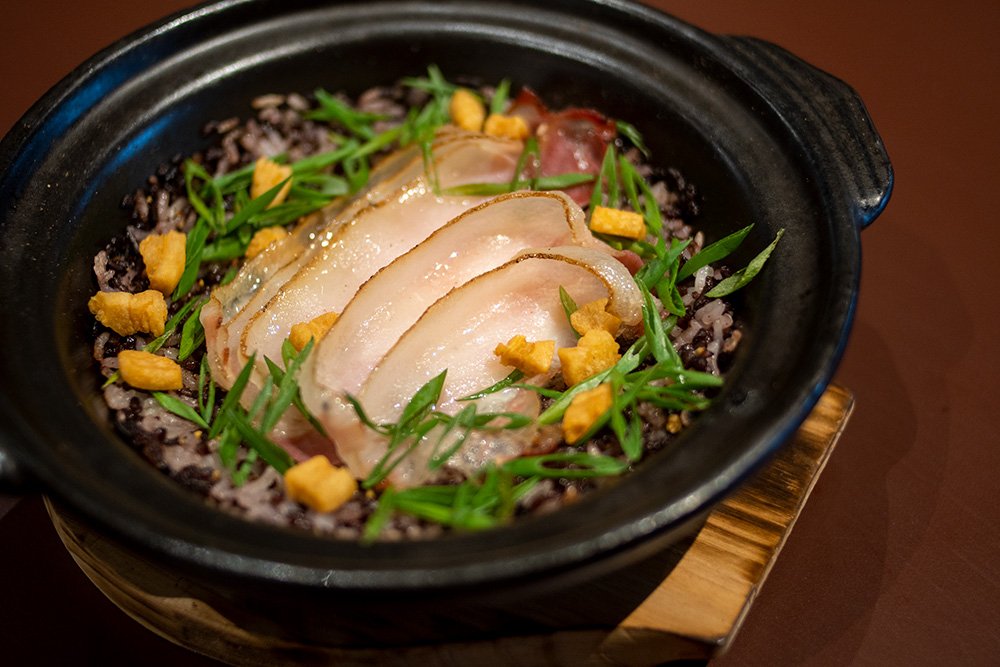 Donabe pot at Bouchon Enfin — dark Sarawak heritage rice topped with sliced smoked pork belly, scallions, and crisp pork puffs.
