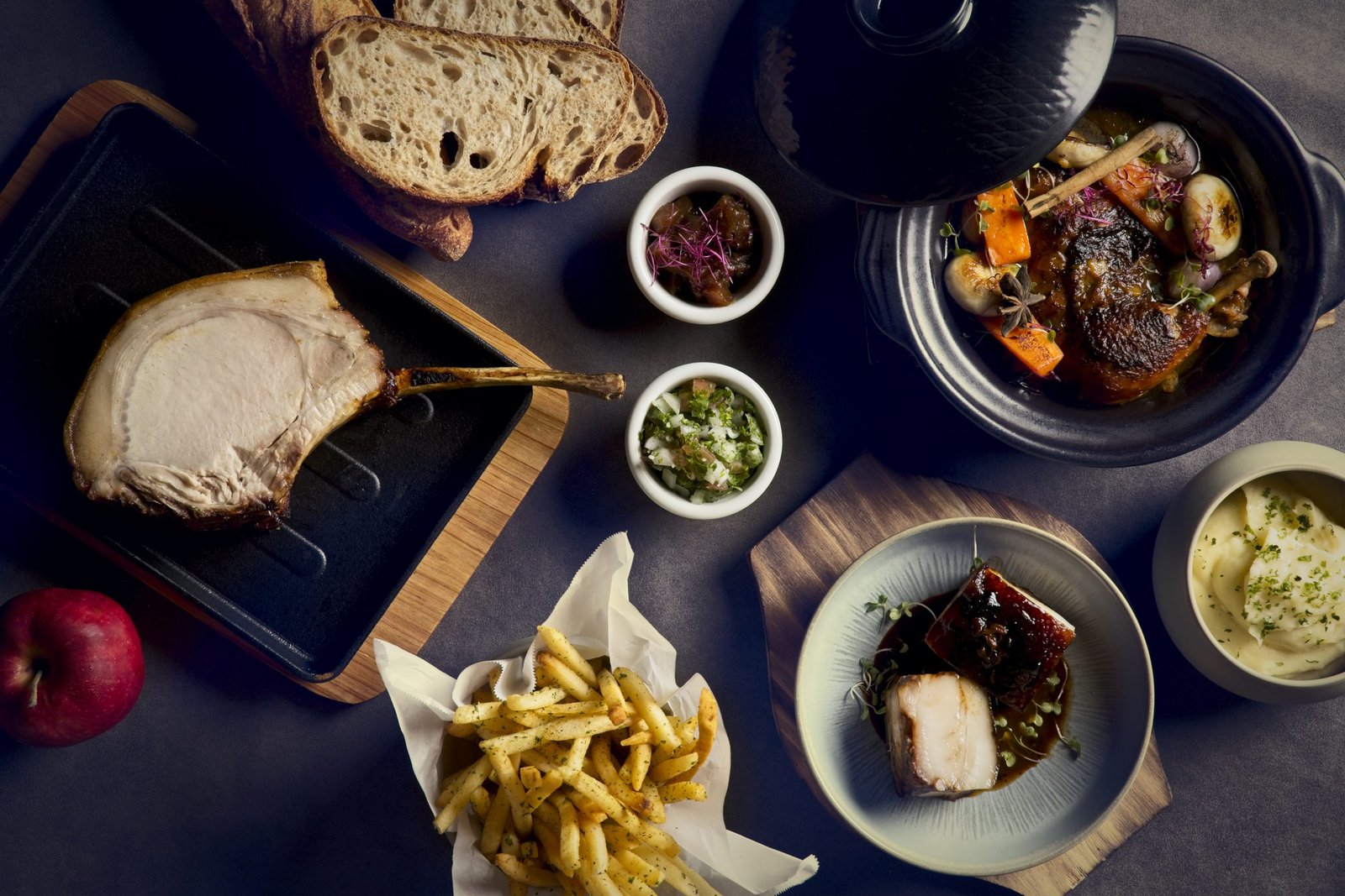 Bouchon Enfin table spread — pork chop on board, sourdough, coq au vin in donabe, glazed pork belly, frites, condiments, mashed potato, all set together.