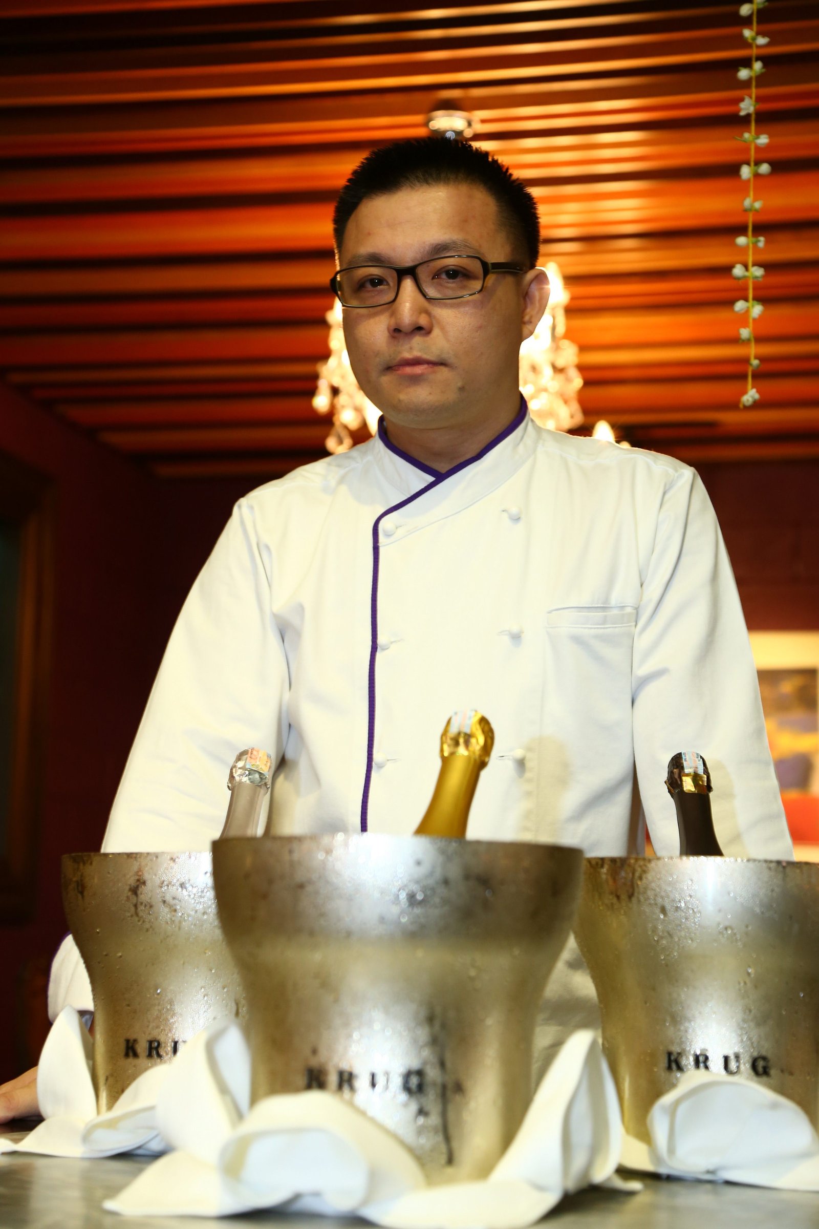 James Won at Brasserie Enfin opening — chef in white jacket with violet trim, behind three Krug ice buckets with bottles, the burgundy room behind.