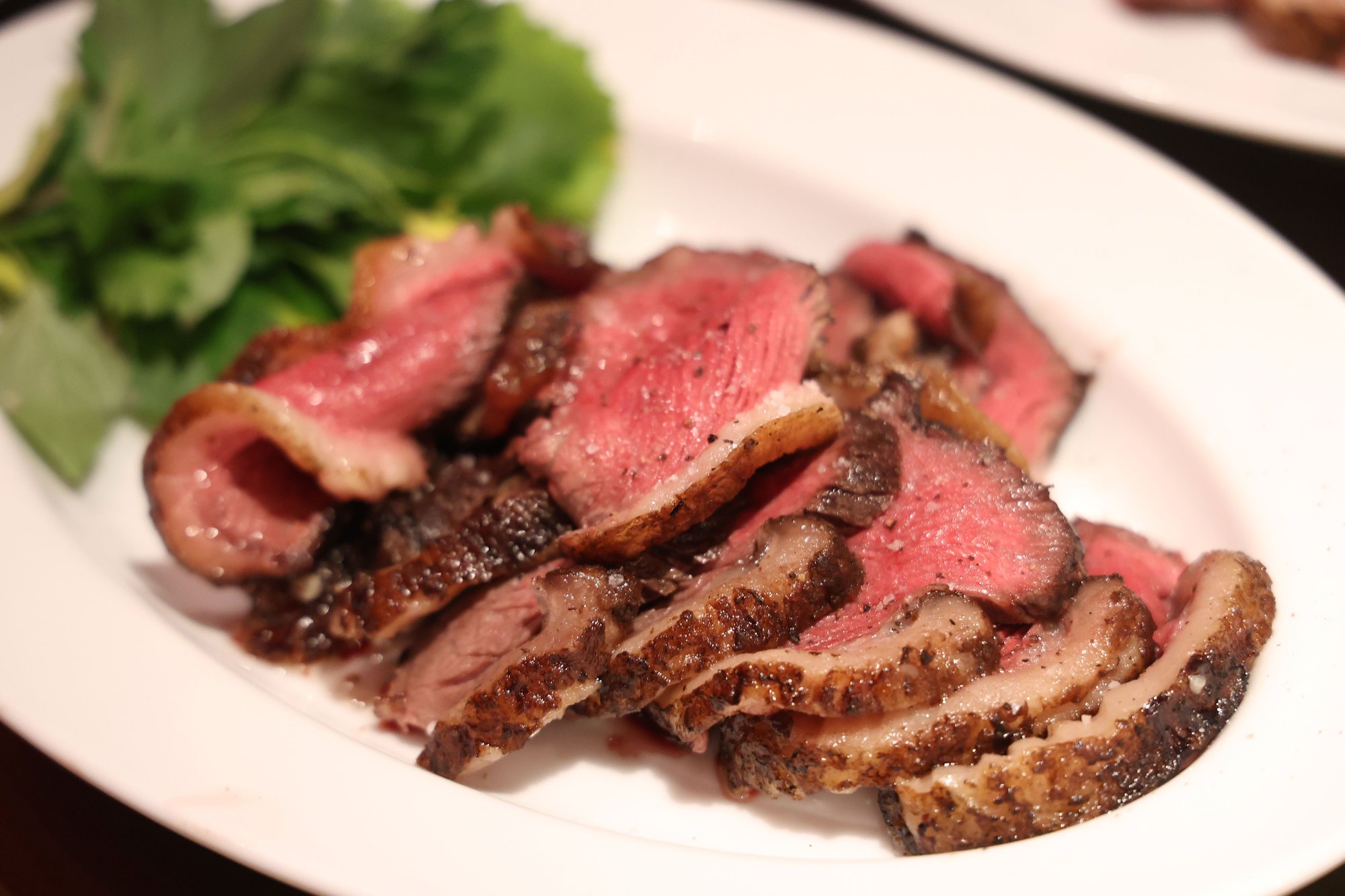 A steak sliced into thick pink pieces on a white plate, with greens.