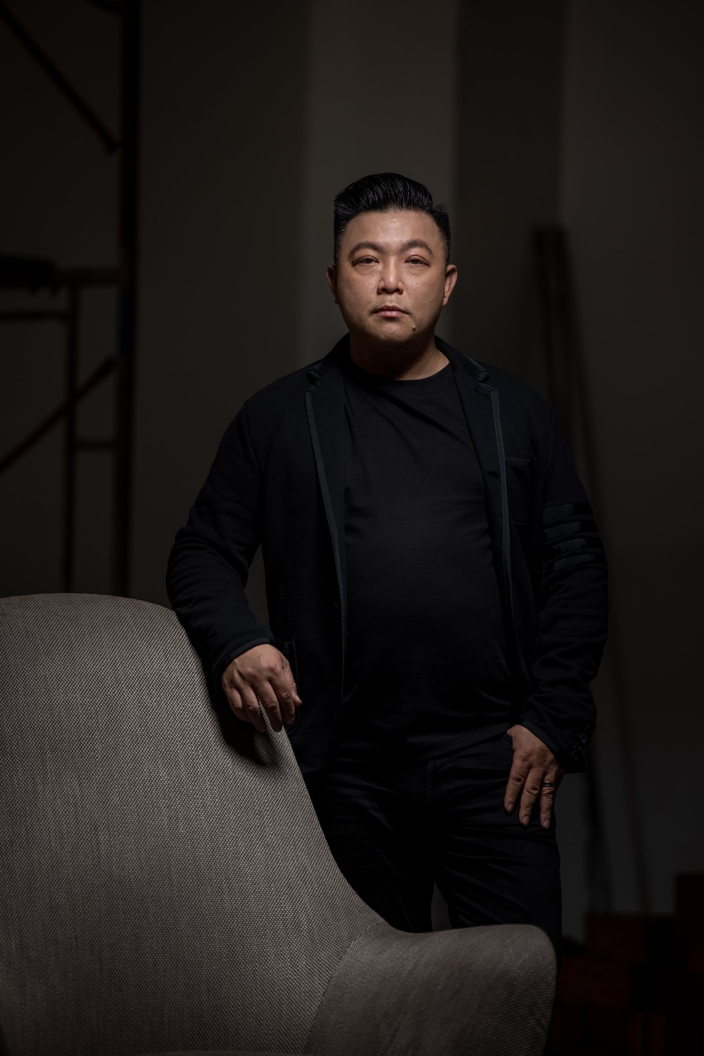 James Won in black, standing beside a grey armchair, one hand resting on its back, against a dark studio wall.