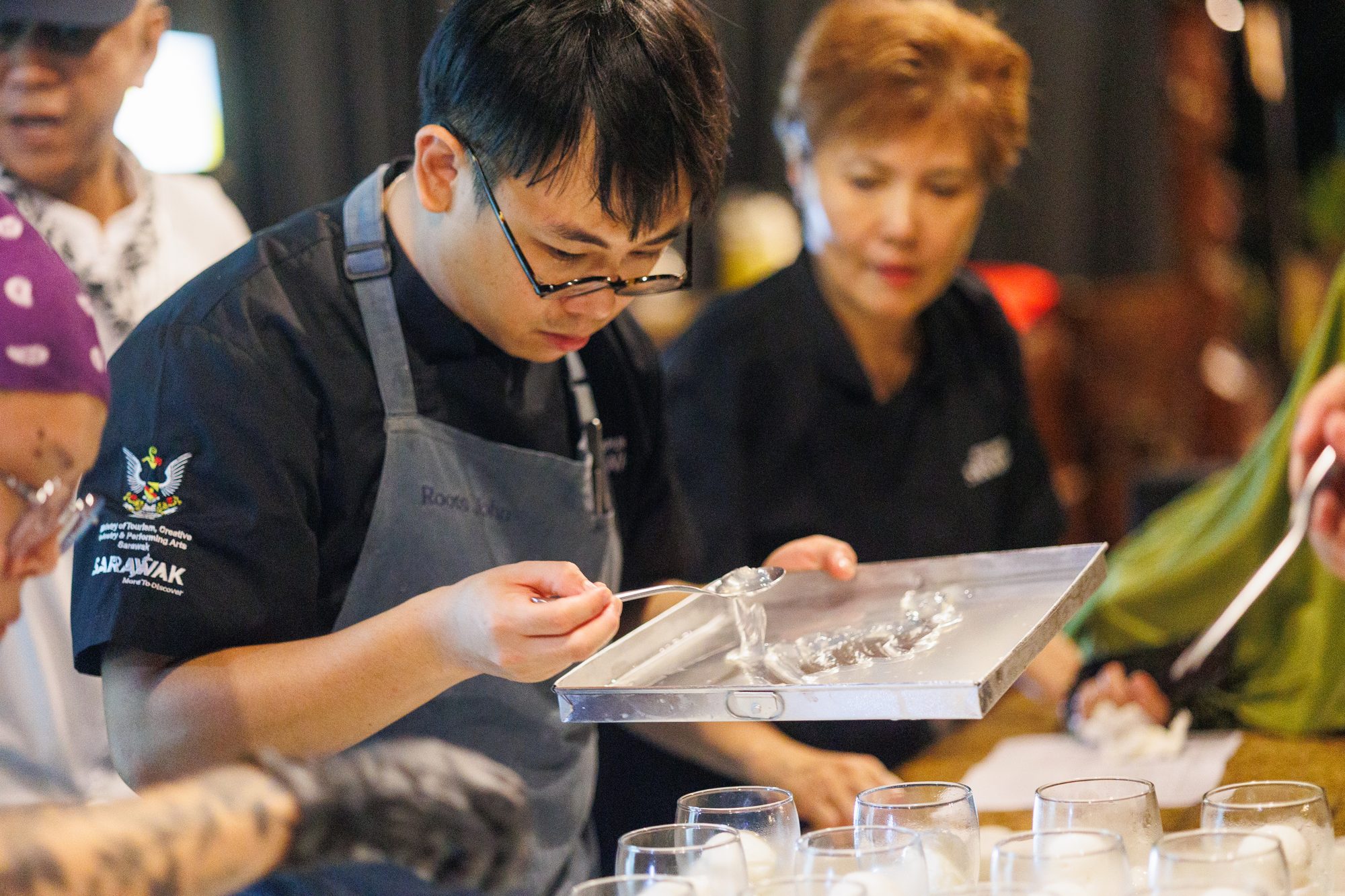 A chef spooning a clear jelly onto a steel tray at the Kuching Finale, glasses lined up for service in front of him